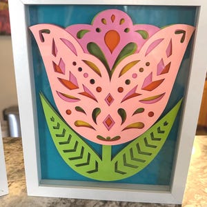 May include: A framed, layered paper art piece featuring a stylized pink tulip with intricate cut-out details. The flower has green leaves and is set against a teal background within a white frame. The artwork is a decorative piece.