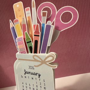 Può includere: Calendario di carta per gennaio 2026, a forma di barattolo pieno di materiale scolastico. Il calendario include un righello, matite, pastelli, forbici e penne. Il barattolo ha un fiocco ed è su un supporto trasparente.