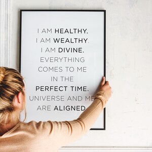 Może przedstawiać: Oprawiony wydruk z czarną obwódką wyświetla tekst "I AM HEALTHY. I AM WEALTHY. I AM DIVINE." Wydruk jest trzymany przez osobę noszącą beżową koszulę z długim rękawem. Tło to biała ściana.