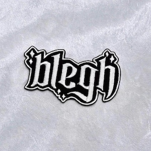 Peut inclure: Écusson brodé noir et blanc avec le mot "blegh" dans une police gothique. Le patch a une bordure blanche et est posé sur un fond de tissu gris. Idéal pour les vêtements ou les accessoires.
