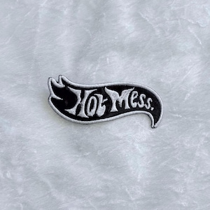 Peut inclure: Écusson brodé noir et blanc avec l'inscription "Hot Mess" dans une police stylisée. L'écusson a la forme d'une bannière incurvée avec une bordure blanche. Il est conçu pour être cousu ou thermocollé sur des vêtements ou accessoires.