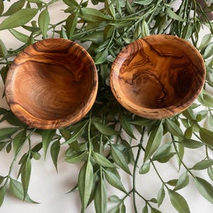 May include: Two round wooden bowls, displaying a rich, warm brown hue and distinctive wood grain patterns. The bowls have a smooth interior and are set against a backdrop of green foliage. These bowls are suitable for serving food or as decorative items.