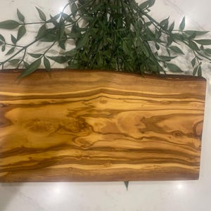 May include: A rectangular olive wood chopping board with a natural edge. The board has a warm, golden-brown colour and prominent wood grain patterns. Green leafy sprigs are arranged above the board.