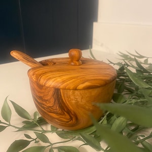 May include: A wooden bowl with a lid and a small spoon. The bowl and spoon are made of wood with a natural grain pattern. The lid has a small knob on top. The bowl is sitting on a surface with green leaves.