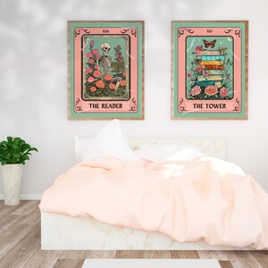 May include: Two framed tarot card prints. The Reader features a skeleton reading among roses, and The Tower shows a stack of books with a butterfly. Both have pink and teal backgrounds with wooden frames. The prints are displayed above a bed.