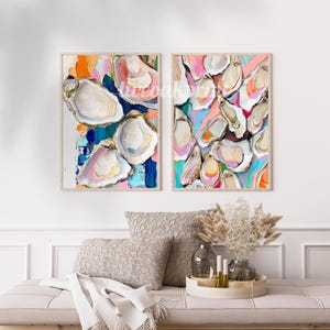 May include: Two framed abstract paintings of oyster shells in shades of pink, blue, yellow, and white. The paintings are set against a white wall with a sofa and decorative plants in the foreground.