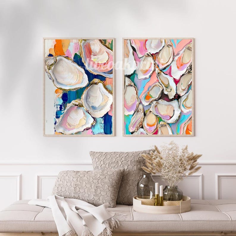 May include: Two framed abstract paintings of oyster shells in shades of pink, blue, yellow, and white. The paintings are set against a white wall with a sofa and decorative plants in the foreground.