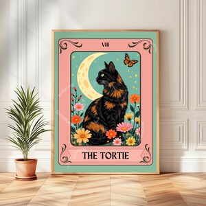 May include: A framed print featuring a black and orange tortoiseshell cat, a crescent moon, and a butterfly. The artwork is set against a teal background with colorful flowers and the text "THE TORTIE" at the bottom. The frame is a light green color.