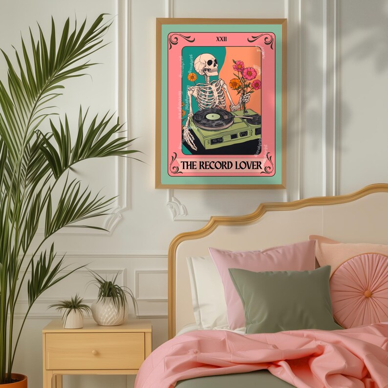Op de afbeelding: Een ingelijste kunstprint met een skelet-dj met een platenspeler en bloemen. Het kunstwerk heeft een roze en teal kleurenschema met de tekst "THE RECORD LOVER" onderaan. De print hangt boven een bed met roze beddengoed.