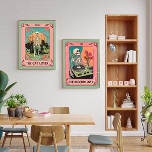 Op de afbeelding: Twee ingelijste kunstdrukken met een vintage tarotkaartontwerp. E&eacute;n toont een skelet met katten, de tekst "THE CAT LOVER". De andere toont een skelet met een platenspeler, met de tekst "THE RECORD LOVER". Beide hebben een roze en teal kleurenschema.