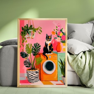May include: A framed art print featuring a colorful illustration of a cat sitting on an orange washing machine. The artwork includes potted plants, flowers, and a pink background. The cat is black, white, and orange.