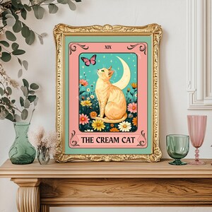 May include: A framed art print featuring a cream-colored cat gazing upwards, set against a backdrop of flowers, a crescent moon, and a butterfly. The text "THE CREAM CAT" is displayed at the bottom, with "XIX" at the top.