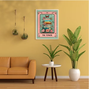 May include: A framed tarot card print featuring a stack of books, flowers, and a butterfly. The card reads "THE TOWER" in a decorative font. The print is displayed on a yellow wall, with a brown leather sofa and plants in the foreground.
