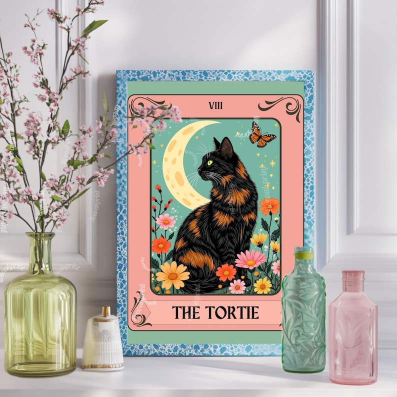 May include: A framed print featuring a black and orange tortoiseshell cat, a crescent moon, and colorful flowers. The text "THE TORTIE" is at the bottom. The print is surrounded by a pink and blue border. Decorative bottles and a vase are in the foreground.
