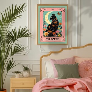 May include: A framed art print featuring a black and orange cat, a crescent moon, and a butterfly. The text "THE TORTIE" is at the bottom. The artwork is set against a pink and teal background, with a gold frame.