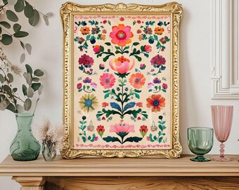 Op de afbeelding: Een ingelijste bloemenprint met een gouden, sierlijke lijst. Het kunstwerk toont een symmetrisch ontwerp van kleurrijke bloemen in tinten roze, oranje en paars op een crèmekleurige achtergrond. De print wordt tentoongesteld op een houten schoorsteenmantel.