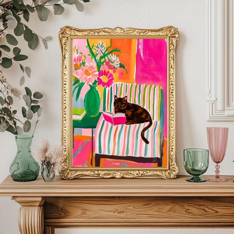Op de afbeelding: Een ingelijste kunstdruk toont een kat die een boek leest op een gestreepte stoel. Het kunstwerk maakt gebruik van een levendig kleurenpalet met roze, oranje en groene tinten. Een gouden lijst omringt de afbeelding, die een vaas met bloemen bevat.