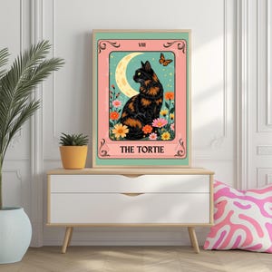 May include: A framed art print featuring a black and orange cat illustration. The artwork has a pink and teal background with a crescent moon, flowers, and a butterfly. The text "THE TORTIE" is at the bottom.