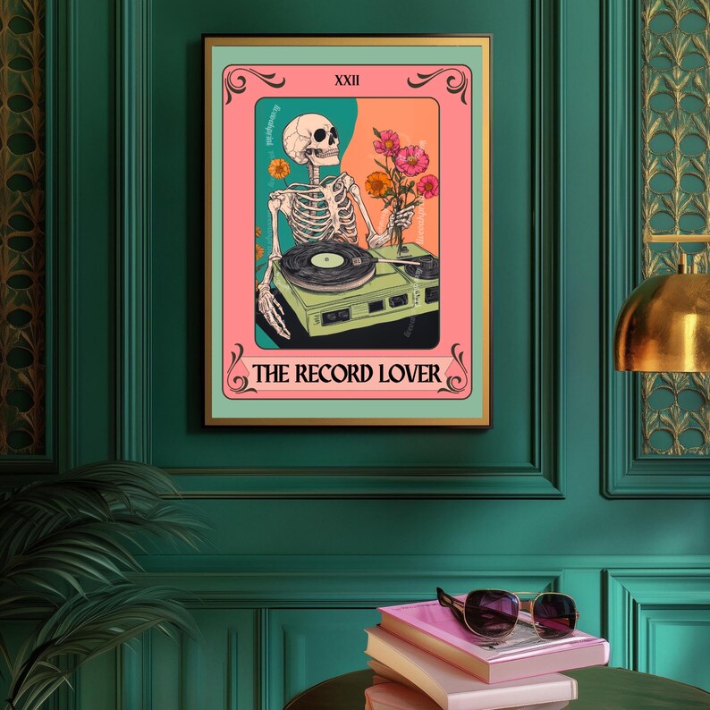 Op de afbeelding: Een ingelijste kunstprint met een skelet-DJ met een platenspeler en bloemen. Het kunstwerk heeft een roze, oranje en groen kleurenschema met de tekst "THE RECORD LOVER". De lijst is goudkleurig.