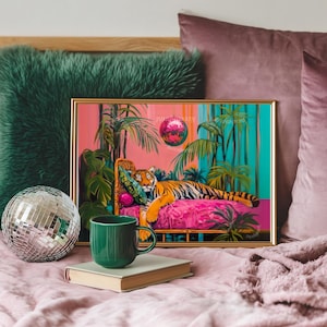 May include: A framed artwork depicts a tiger lounging on a pink chaise lounge, surrounded by tropical plants. A green mug sits on a book in front of the artwork. A disco ball and green pillow are also in the scene. The artwork has a gold frame.