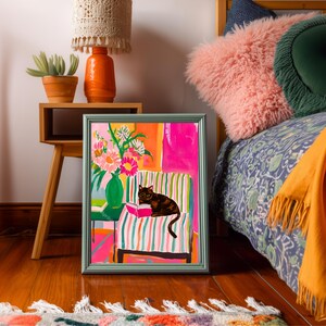 Op de afbeelding: Een ingelijst schilderij met een zwarte kat die een boek leest op een gestreepte stoel. Het kunstwerk heeft een levendig kleurenpalet met roze, oranje en groene tinten. Een vaas met bloemen en een bijzettafel met een lamp zijn ook zichtbaar.