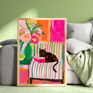 Op de afbeelding: Een ingelijste kunstdruk toont een bruine kat die een boek leest op een gestreepte stoel. Een groene vaas met roze en witte bloemen staat op een groene tafel. De achtergrond heeft oranje en roze blokken.