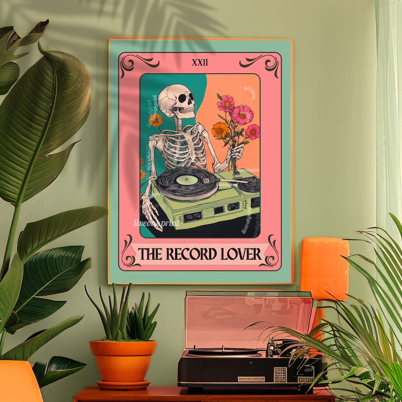 Op de afbeelding: Een ingelijste kunstdruk met een skelet-DJ, platenspeler en bloemen. Het kunstwerk heeft een roze en oranje achtergrond met de tekst "The Record Lover". De lijst is lichtgroen. Het thema is muziek.