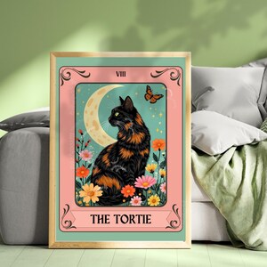 May include: A framed print featuring a black and orange tortoiseshell cat, labeled "THE TORTIE." The artwork includes a crescent moon, butterfly, and colorful flowers. The background is a teal color, and the frame is gold.