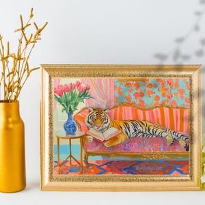 Puede incluir: Una pintura de un tigre leyendo un libro mientras descansa en un sofá a rayas rosa y naranja. El tigre es naranja y negro con ojos verdes. El sofá está en una habitación con papel tapiz floral rosa y azul. La pintura está enmarcada en oro.