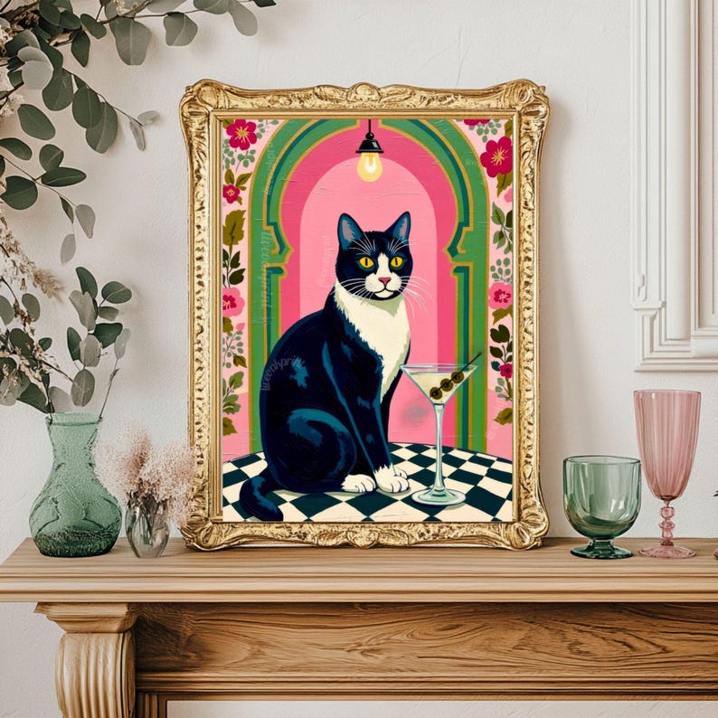 May include: A framed art print featuring a black and white cat with yellow eyes, sitting next to a martini glass with olives. The background has pink and green arches with floral accents, and the frame is gold.