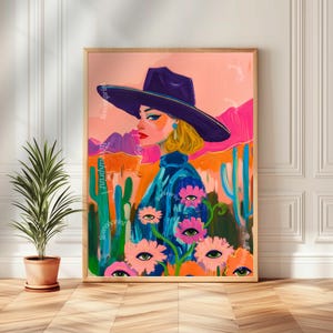 May include: A framed art print featuring a stylized portrait of a woman wearing a wide-brimmed hat. The artwork has a desert landscape background with cacti and flowers with eye designs. The color palette includes pink, orange, blue, and purple.