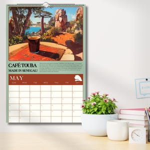 Puede incluir: Un calendario de pared con una ilustraci&oacute;n vibrante de un vaso de caf&eacute; sobre una mesa con un fondo esc&eacute;nico. El calendario est&aacute; etiquetado como "CAF&Eacute; TOUBA MADE IN SENEGAL!" y muestra el mes de mayo. El calendario mide aproximadamente 30 cm de ancho.