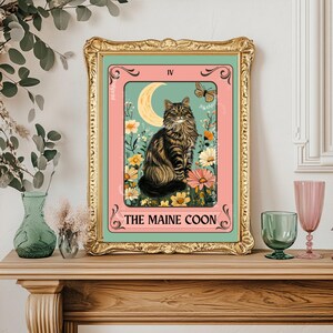 May include: A framed art print featuring a Maine Coon cat illustration. The artwork has a gold frame and a pink and teal background with floral and moon elements. The text "THE MAINE COON" is at the bottom.