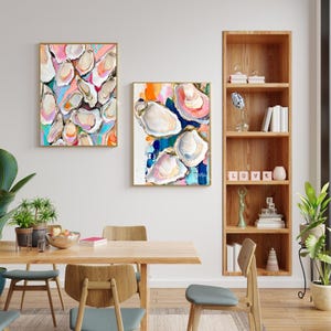 May include: Two abstract paintings of oyster shells in a variety of colours, including pink, blue, green, and yellow. The paintings are framed in gold and hung on a white wall above a wooden table with chairs.