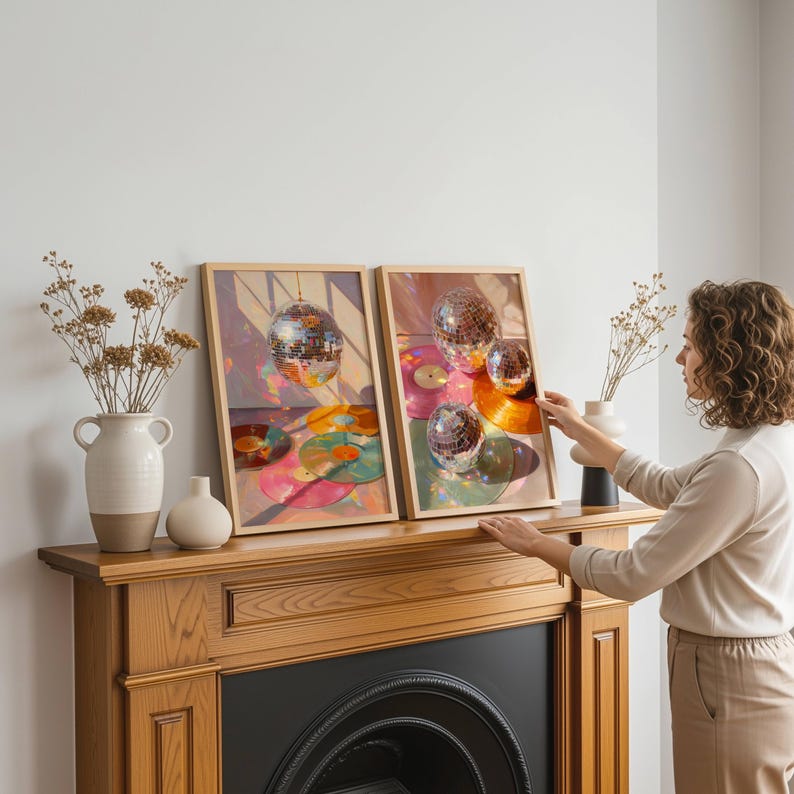 K&ouml;nnte beinhalten: Zwei gerahmte Kunstdrucke mit Discokugeln und Schallplatten in leuchtenden Farben. Die Drucke werden auf einem h&ouml;lzernen Kaminsims ausgestellt, zusammen mit Vasen mit getrockneten Blumen. Eine Person richtet einen der Drucke aus.