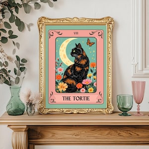 May include: A framed art print featuring a black and orange cat, a crescent moon, and a butterfly. The artwork is set against a pink background with floral accents and the text "THE TORTIE". The frame is gold.