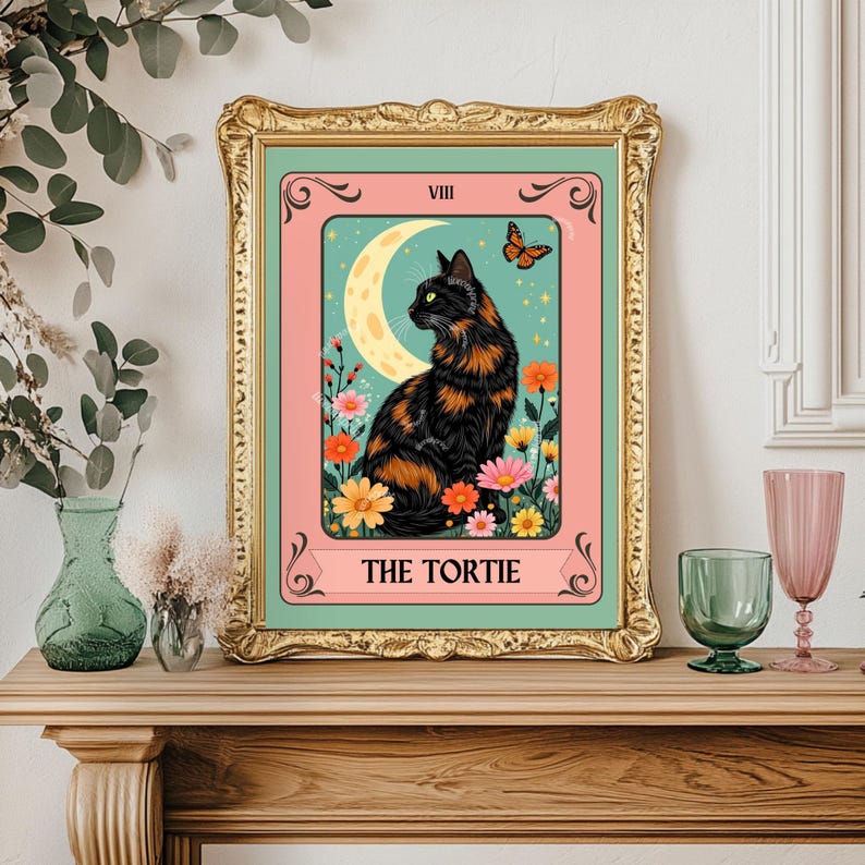 May include: A framed art print featuring a black and orange cat, a crescent moon, and a butterfly. The artwork is set against a pink background with floral accents and the text "THE TORTIE". The frame is gold.