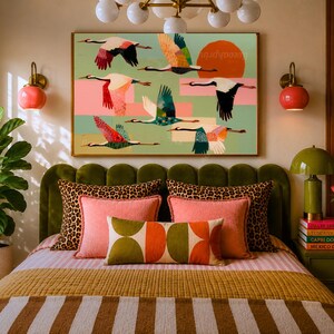 May include: A rectangular wall art print featuring a flock of stylized cranes in flight against a light green background with a large orange sun, layered with pink and yellow rectangular shapes, displayed in a thin gold frame above a bed.