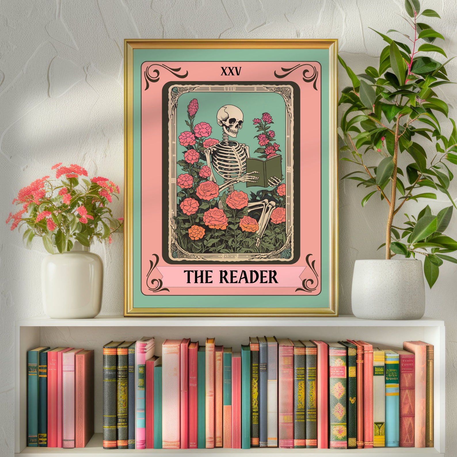 A print in a gold frame has a layer of teal, a layer of pink with whimsical designs and XXV / THE READER on the top and bottom respectively, and the center has a skeleton reading a green book surrounded by pink roses with green leaves.
