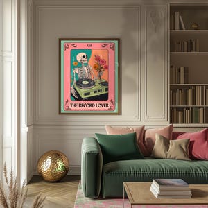 Op de afbeelding: Een ingelijste kunstdruk met een skelet-dj met een platenspeler en bloemen. Het kunstwerk heeft een roze en teal kleurenschema met de tekst "The Record Lover" onderaan. De print wordt in een woonkamer getoond.