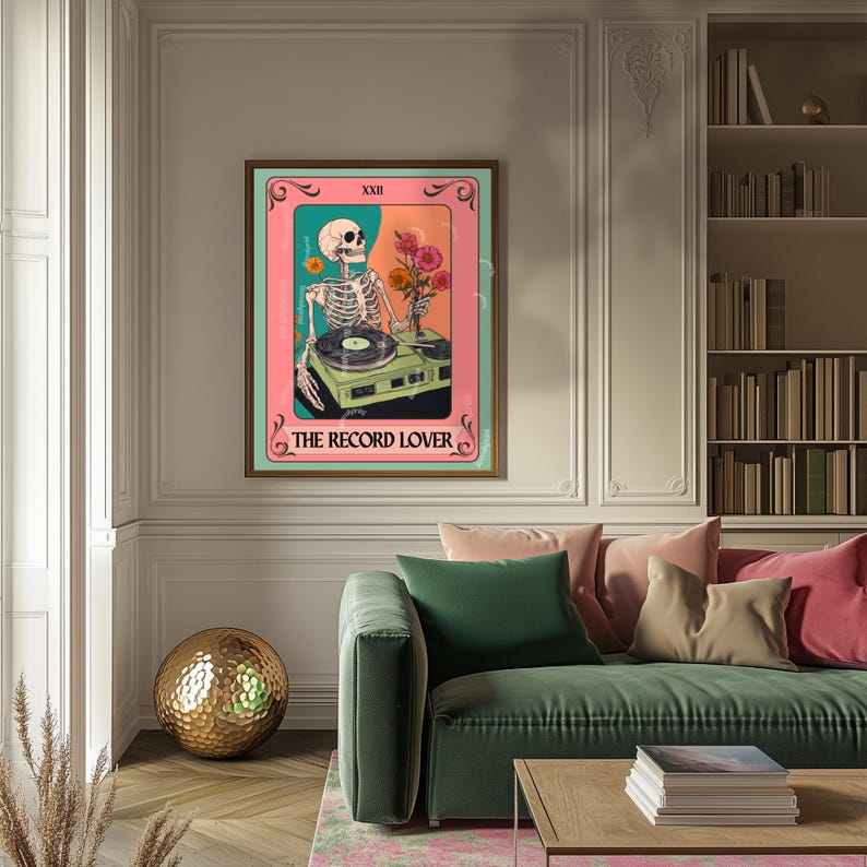 Op de afbeelding: Een ingelijste kunstdruk met een skelet-dj met een platenspeler en bloemen. Het kunstwerk heeft een roze en teal kleurenschema met de tekst "The Record Lover" onderaan. De print wordt in een woonkamer getoond.