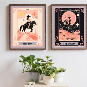 May include: Two framed tarot card prints, one depicting The Sun and the other The Moon. Both cards feature a woman riding a horse in a desert landscape. The Sun card has a yellow sun and a pink sky, while the Moon card has a black sky with a crescent moon and stars.