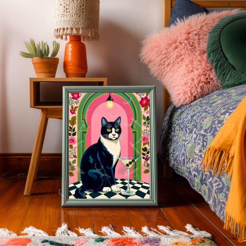 May include: A framed art print featuring a tuxedo cat sitting in front of a martini glass. The cat is black and white with yellow eyes. The background is pink with green archways and floral accents. The print is displayed on a wooden floor.