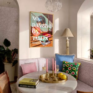 Op de afbeelding: Interieur van een kamer met een ronde marmeren tafel. Een gouden dienblad bevat kaarsen en citroenen. Een ingelijste print met de tekst "NO DANCING ON TABLES" hangt aan de muur. Kussens in roze, blauw en groen voegen kleur toe.