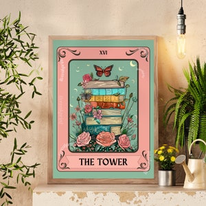 May include: A framed tarot card print featuring a stack of books adorned with flowers and butterflies. The card reads "THE TOWER" at the bottom, with "XVI" at the top. The color palette includes pink, teal, and various book colors.