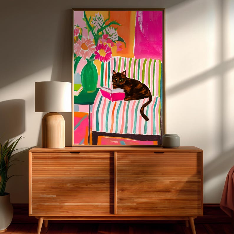 Op de afbeelding: Een ingelijste kunstdruk met een kleurrijke interieursc&egrave;ne. Een bruine kat leest een boek op een gestreepte bank. Een vaas met bloemen staat op een tafel. Het kunstwerk heeft een levendig kleurenpalet van roze, groen en oranje.