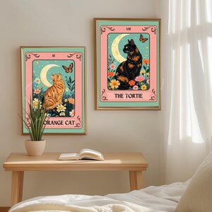 May include: Two framed art prints featuring cat illustrations in a tarot card style. One shows an orange tabby cat, the other a black and orange cat. Both have a crescent moon, flowers, and butterflies.