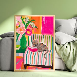 May include: A framed art print featuring a cat reading a book on a striped couch. The artwork has a vibrant color palette with pink, orange, and green hues. A green vase with flowers sits on a table next to the cat.