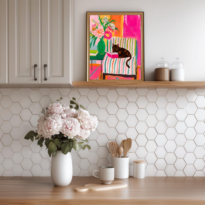 Op de afbeelding: Een ingelijste kunstafdruk toont een zwarte kat die op een gestreepte stoel ligt en een boek leest. Het kunstwerk gebruikt levendige kleuren, waaronder roze, oranje en groen, met een vaas met bloemen. De afdruk wordt boven een houten plank in een keukenomgeving weergegeven.