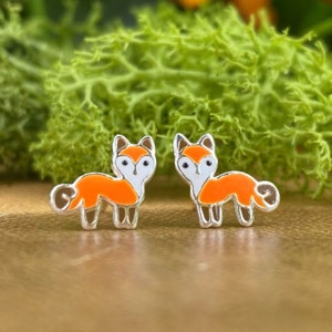 May include: A pair of fox-shaped stud earrings. Each earring features a silver-colored outline with a white face, orange body, and brown tail. The earrings are set against a blurred green and brown background.